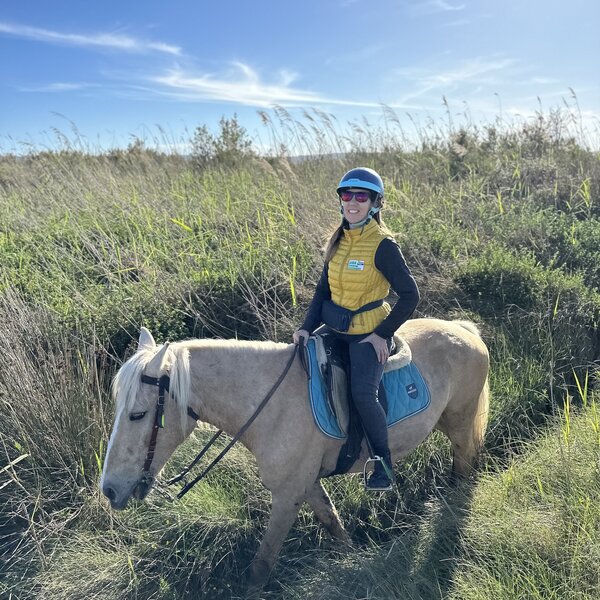 Frau mit Helm und gelber Jacke reitet bei Sonnenschein auf einem Pferd im Gras