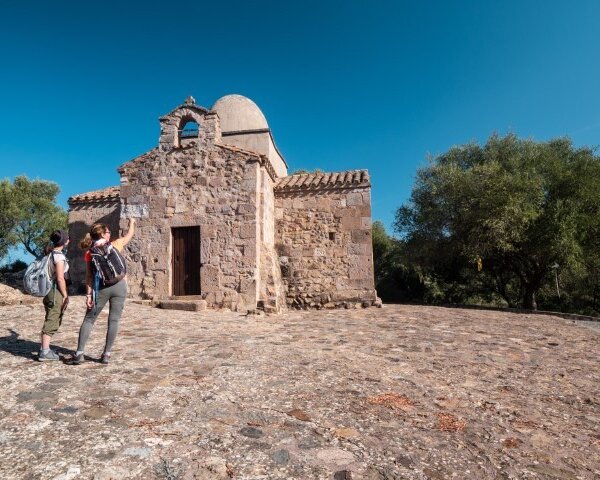 Two people observe a small stone church in a sunny landscape.