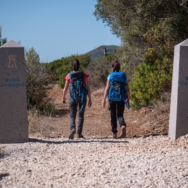 Two hikers with backpacks walk on a trail between two stones reading Buon Cammino.