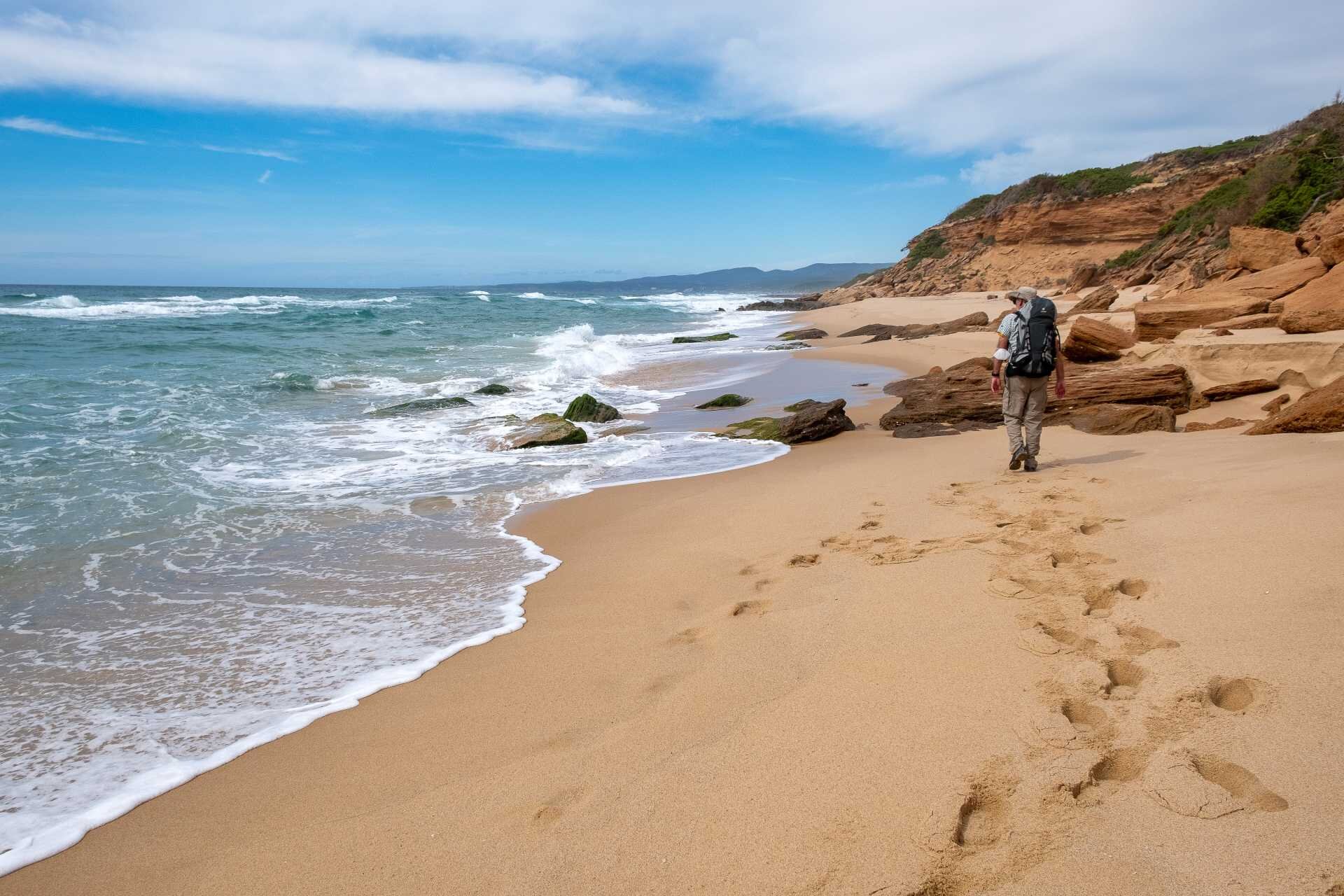 Hiker with backpack walking on sandy beach near rocks and rough sea
