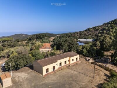 Vue aérienne d'un bâtiment rural entouré d'arbres et de collines sous un ciel clair.