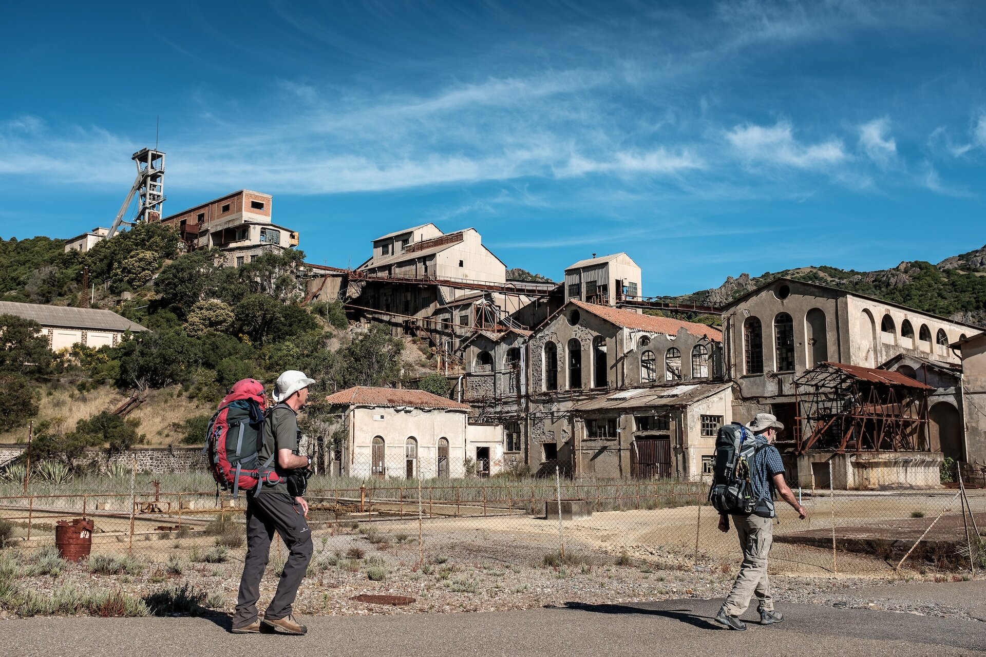 Deux randonneurs avec sacs à dos marchent près de vieux bâtiments industriels abandonnés.