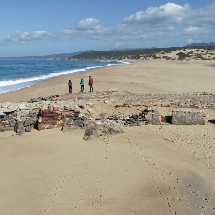 Tre persone camminano vicino a rovine su una spiaggia sabbiosa e deserta.