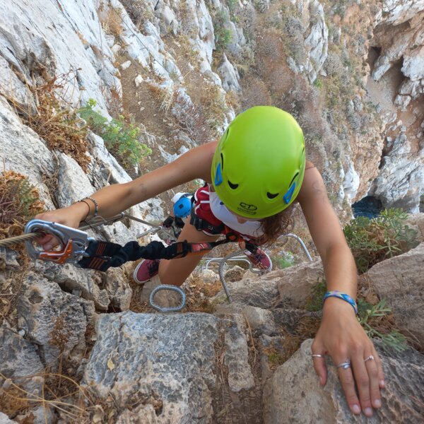 Person wearing green helmet climbing rocky wall with safety harness and carabiners.