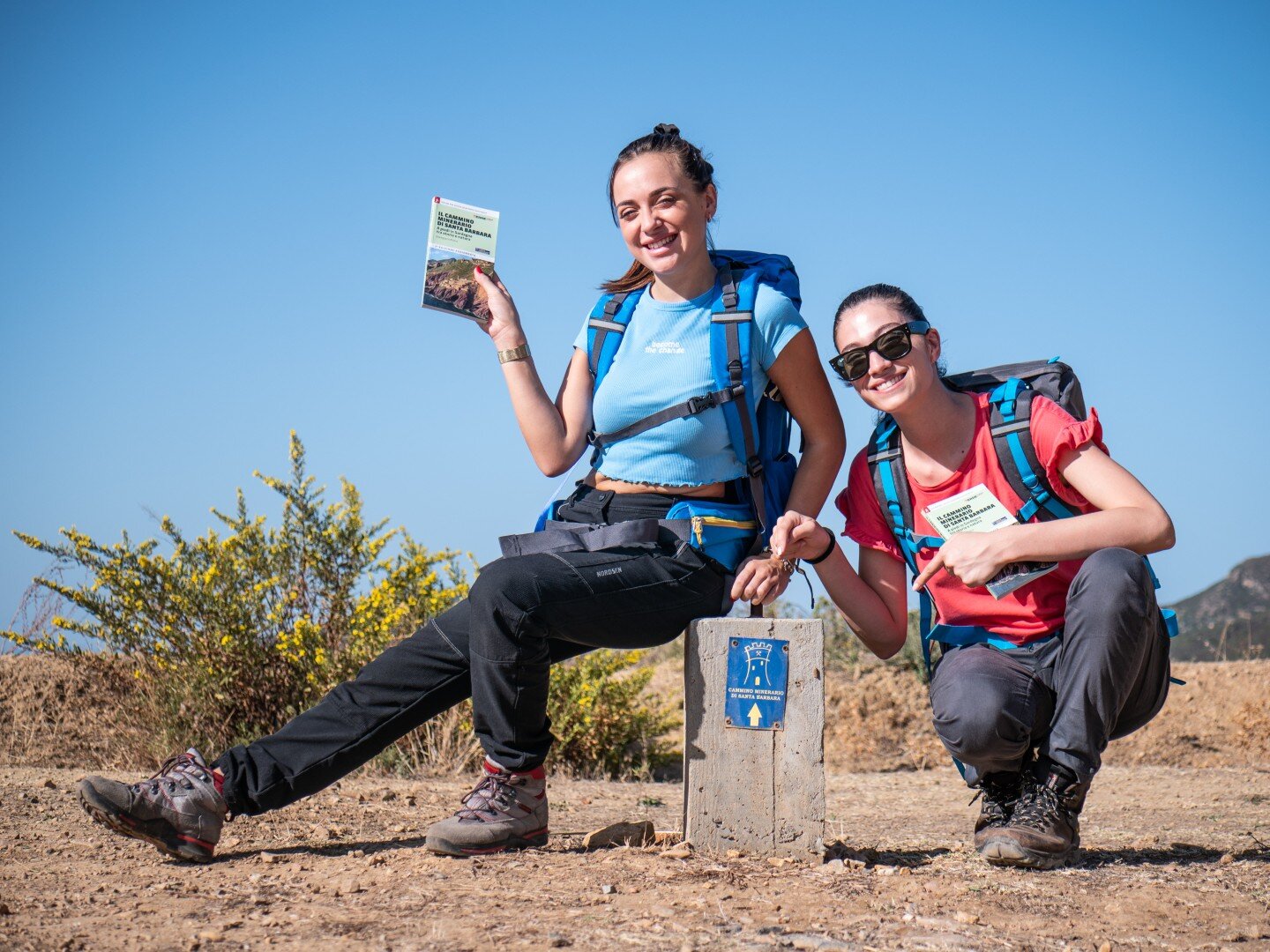Two women with hiking backpacks smiling near a hiking trail marker.