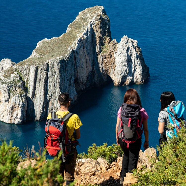 Drei Wanderer betrachten eine große Felsformation über dem blauen Meer.